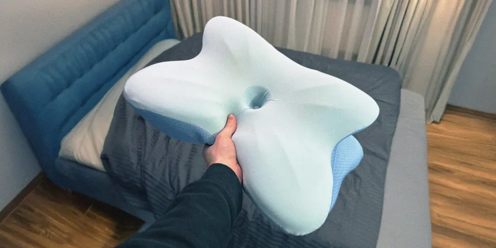The Derila ERGO pillow held in one hand above a bed, showing its butterfly outline and wing sections